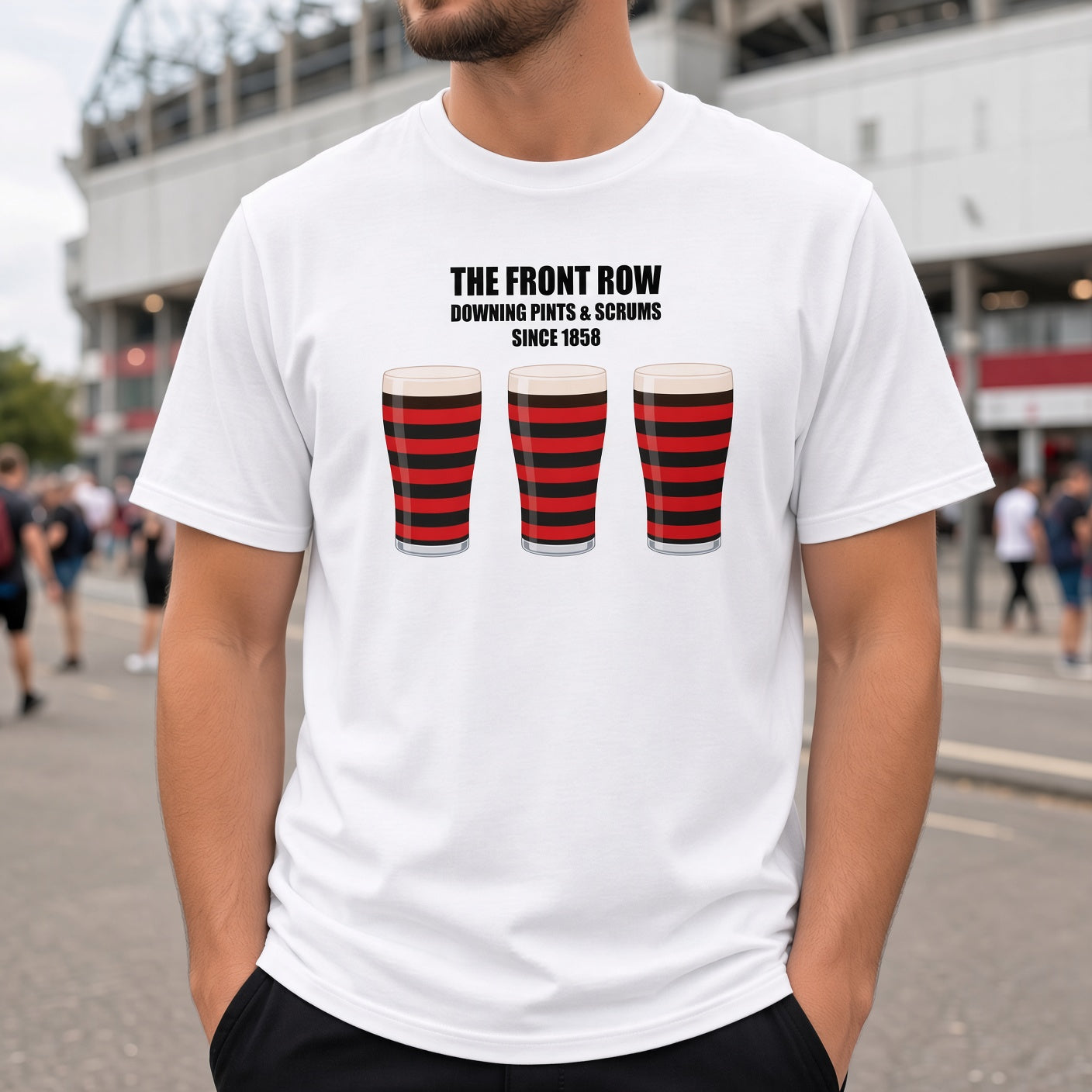 Male model wearing a white Blackheath rugby t-shirt that reads “The Front Row – Downing Pints & Scrums Since 1858” with three red and black pint graphics, photographed on a match day street scene