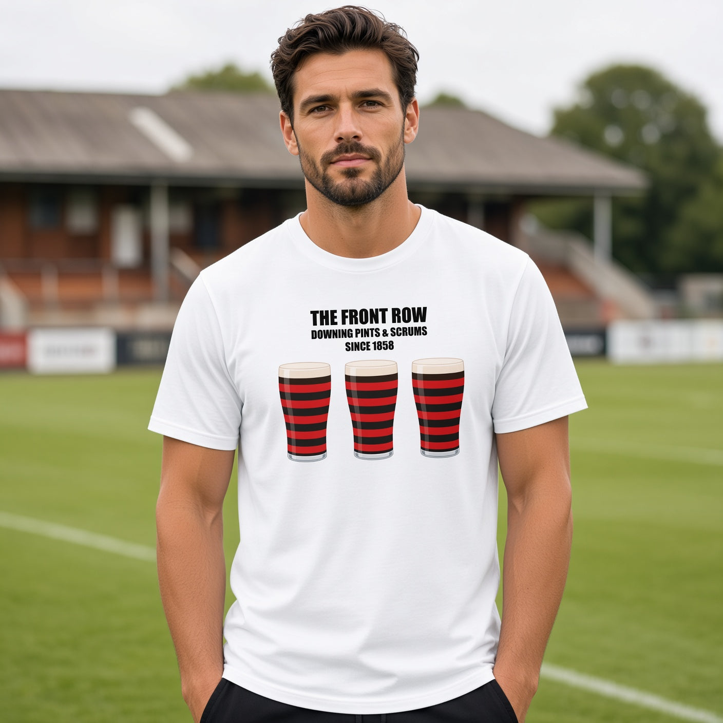 Male model wearing a white Blackheath rugby t-shirt reading “The Front Row - Downing Pints & Scrums Since 1858” with three red and black pint graphics, photographed at a rugby ground for match day supporters