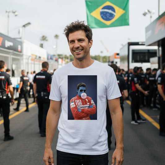 Smiling man wearing TheTeeShop’s Blue Ayrton Bubble Gum T-Shirt from the Paddock Collection, standing in a bustling motorsport paddock with a Brazilian flag in the background, showcasing unique racing-inspired streetwear.