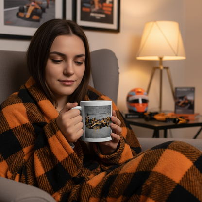A female McLaren fan wrapped in a blanket, sipping from the Papaya Series: Pit Stop Graffiti Mug. A warm, inviting moment surrounded by F1 and McLaren memorabilia.