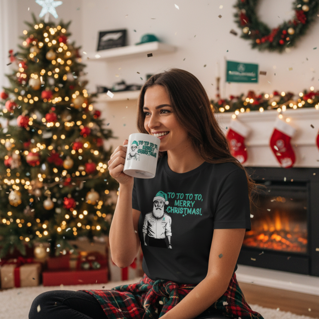 Female model in a Christmas setting, wearing black Toto Wolff Merry Christmas T-Shirt and holding the matching mug, with Mercedes-AMG F1 themed decor and a holiday tree.