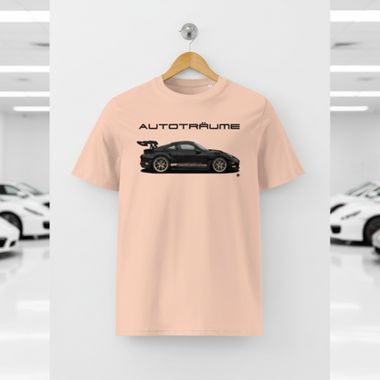 Soft peach AUTOTRÄUME GT3 T‑shirt with a black Porsche‑style car and gold highlights illustration hanging on a premium wooden hanger and silver wall peg against a central white wall in a bright sports car gallery, with a glossy white floor and blurred white and silver sports cars running down both sides.