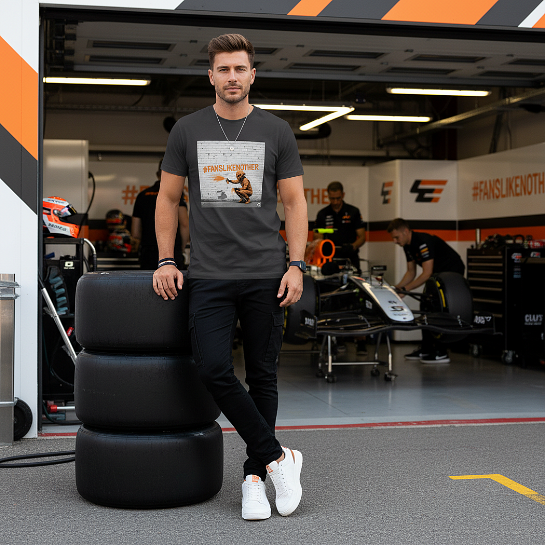Male model wearing the anthracite organic cotton PAPAYA Fans Like No Other Graffiti Art F1 T-Shirt with #FANSLIKENOOTHER design, standing with arms relaxed by his sides in a full-frame motorsport paddock environment, tee clean and design fully visible.
