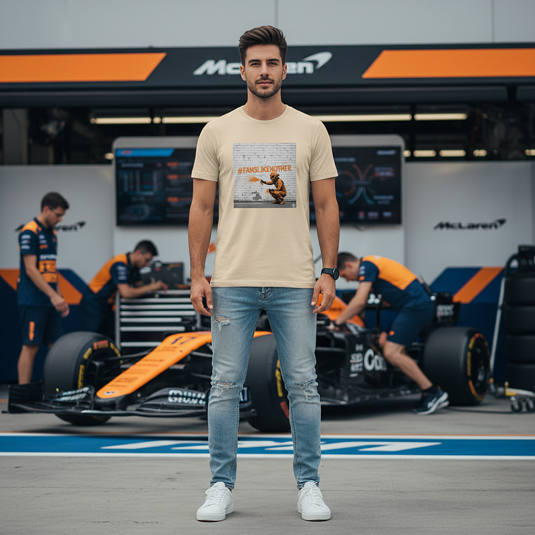Male model wearing the desert dust organic cotton PAPAYA Fans Like No Other Graffiti Art F1 T-Shirt with #FANSLIKENOOTHER design, standing with arms relaxed by his sides in a full-frame motorsport paddock environment, tee clean and design fully visible.