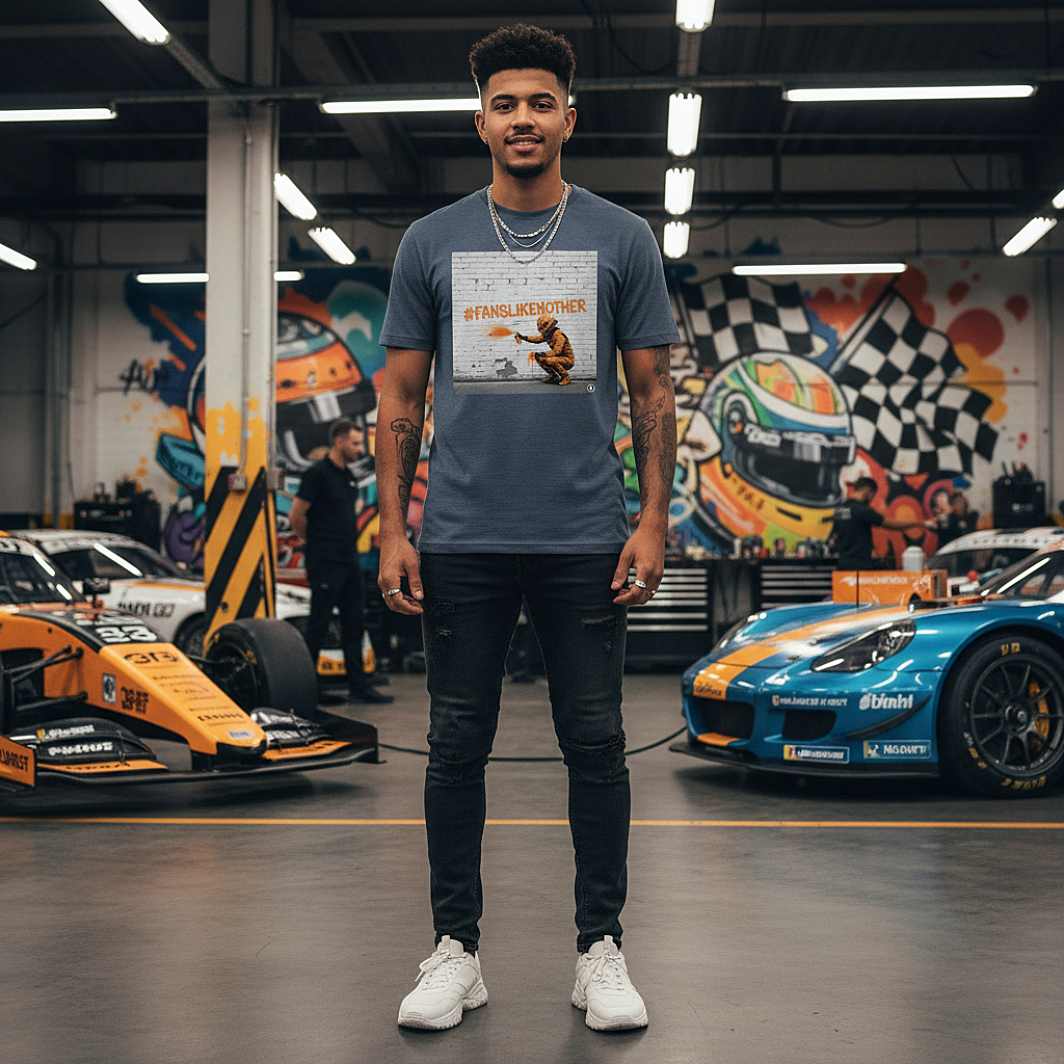 Male model wearing the dark heather blue organic cotton PAPAYA Fans Like No Other Graffiti Art T-Shirt with #FANSLIKENOOTHER design, standing with arms relaxed by his sides in a full-frame motorsport paddock environment, tee clean and design fully visible.