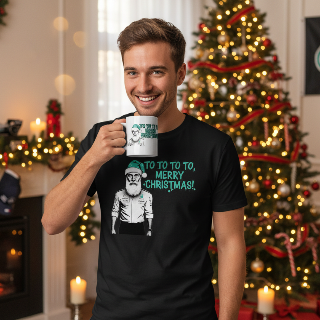 Male model in a festive Christmas setting wearing the Toto Wolff Merry Christmas T-Shirt (white or black) and holding the official Toto Wolff Merry Christmas mug, surrounded by Mercedes-AMG F1 memorabilia and holiday decor.