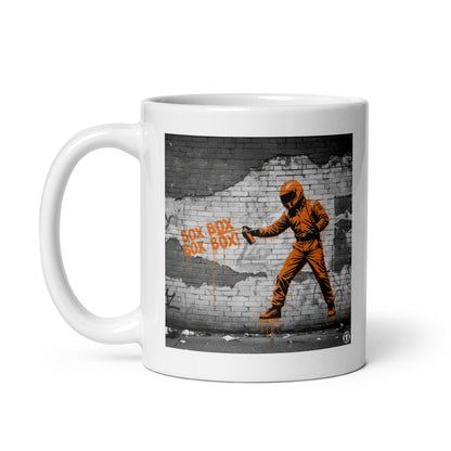 White ceramic 11oz mug featuring a street art illustration of a racing driver in a papaya suit spray-painting “BOX BOX BOX BOX!” in orange on a distressed brick wall, inspired by McLaren F1 pit stops.
