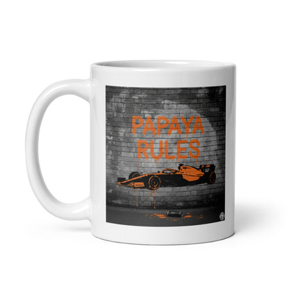 White ceramic 11oz mug featuring a street art illustration of a papaya orange McLaren F1 car and “PAPAYA RULES” graffiti on a dark brick wall.