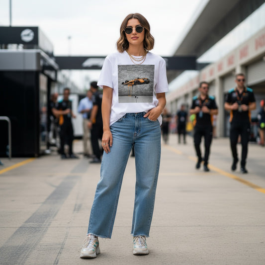 Young woman wearing TheTeeShop’s Papaya Series Graffiti Racer T-Shirt, featuring a graffiti-style papaya orange F1 car, standing in the Formula 1 paddock with team members and garages in the background, showcasing bold McLaren-inspired motorsport streetwear.