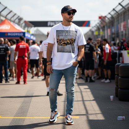Man wearing TheTeeShop’s Papaya Series Pit Stop Graffiti T-Shirt, standing on a busy Formula 1 paddock with team members and fans in the background, showcasing bold McLaren-inspired street art motorsport apparel.