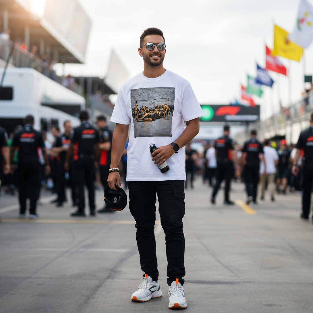 Man wearing TheTeeShop’s Papaya Series Pit Stop Graffiti T-Shirt, walking trackside at a Formula 1 event with team members and fans in the background, showcasing bold McLaren-inspired street art motorsport apparel.
