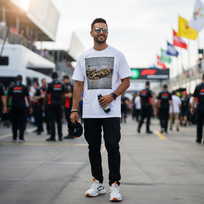 Man wearing TheTeeShop’s Papaya Series Pit Stop Graffiti T-Shirt, walking trackside at a Formula 1 event with team members and fans in the background, showcasing bold McLaren-inspired street art motorsport apparel.
