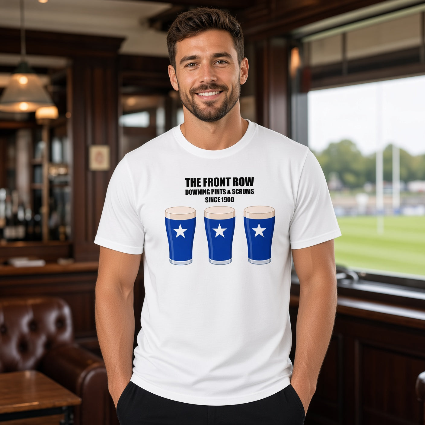 Male model wearing a white St Mary’s rugby t-shirt reading “The Front Row - Downing Pints & Scrums Since 1900” with three blue pint graphics featuring a white star, photographed inside a rugby clubhouse/pub setting
