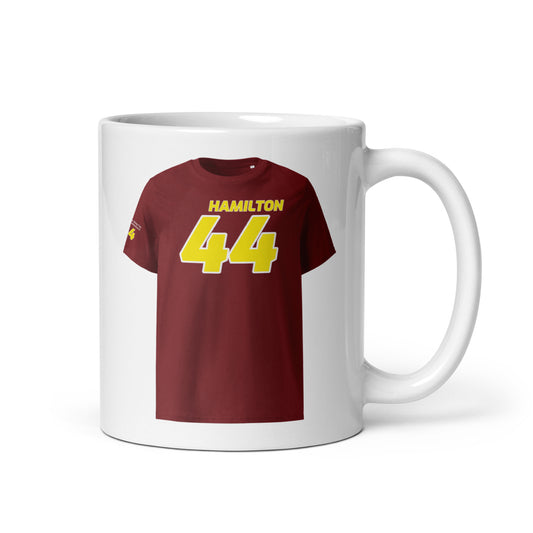 A white ceramic TeeMug© featuring a yellow design with "HAMILTON" in bold letters and the number "44". The mug is held by a hand on the right side.