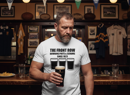 Veteran rugby prop in his 40s wearing a white front row rugby t-shirt with "The Front Row Downing Pints & Scrums Since 1823" slogan and three pint graphics, standing at a traditional UK rugby club bar with memorabilia and trophies displayed behind him.
