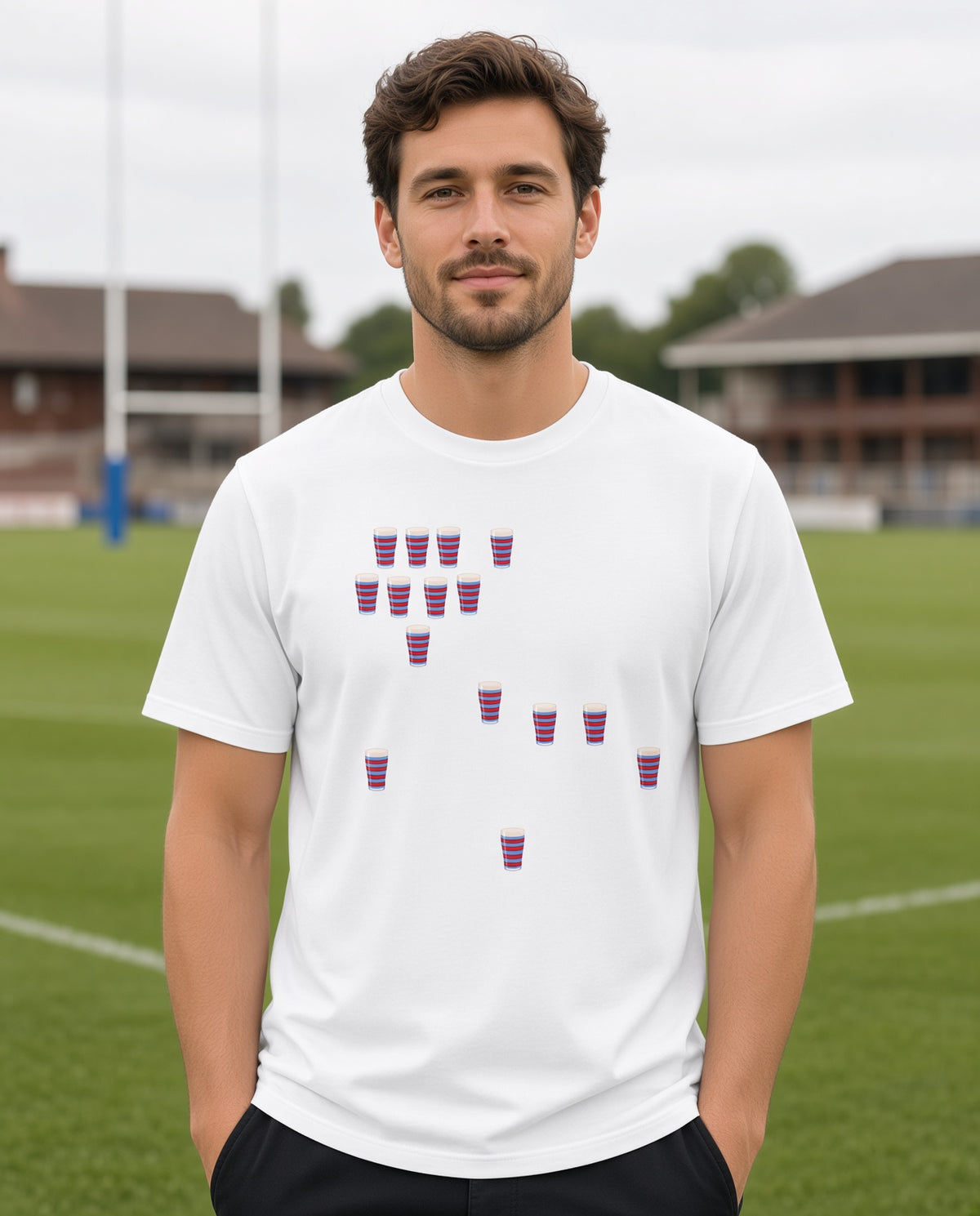 Male model wearing a white vintage Blackheath Team Pints rugby t-shirt with scattered red and light blue striped pint graphics, photographed on a rugby pitch for match day supporters in South East London