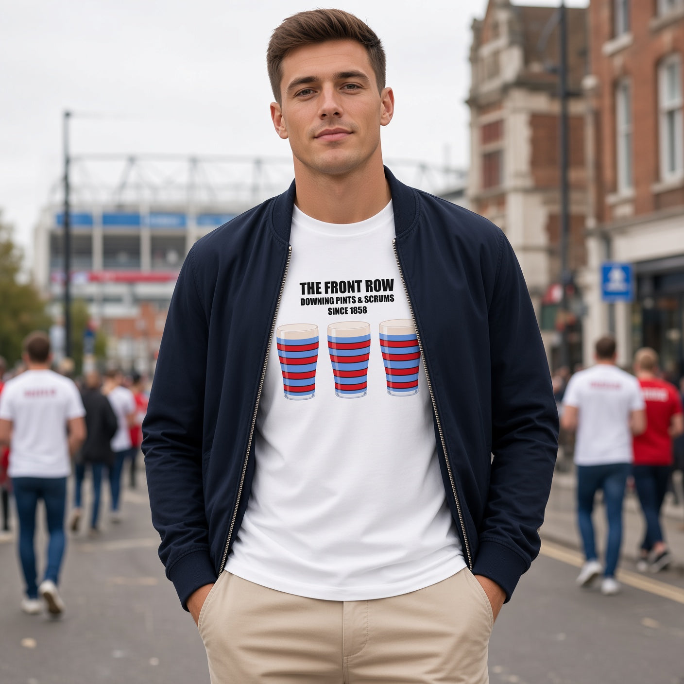 Male model wearing a white vintage Blackheath rugby t-shirt reading “The Front Row – Downing Pints & Scrums Since 1858” with three red and light blue striped pint graphics, photographed in a match day crowd setting