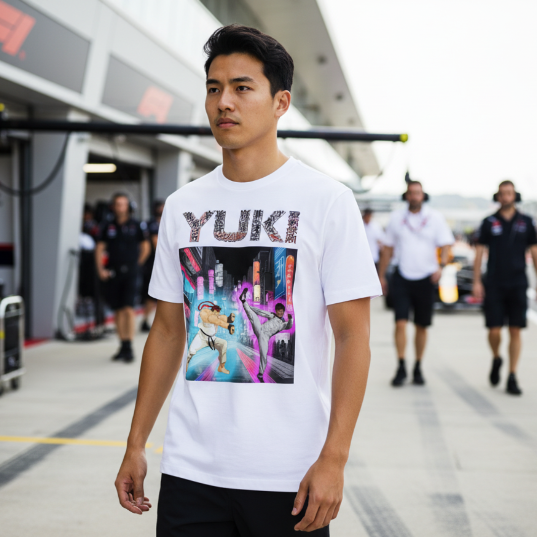 Male motorsport fan in an F1 paddock wearing a white YUKI martial arts T‑shirt, turned slightly to show the neon city fight scene graphic with garages blurred behind