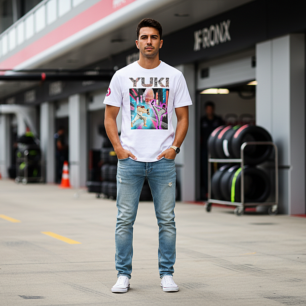 Male motorsport fan in an F1 paddock wearing a white YUKI martial arts T‑shirt with neon city fight scene graphic and YUKI text on the front, garages and pit lane blurred behind.