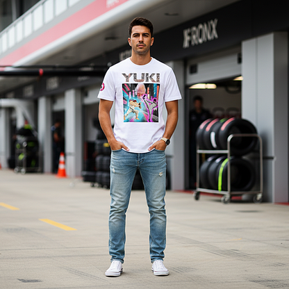 Male motorsport fan in an F1 paddock wearing a white YUKI martial arts T‑shirt with neon city fight scene graphic and YUKI text on the front, garages and pit lane blurred behind.